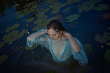 Narin mavi giysiler giymiş bir kadın etrafını saran nilüferlerle çevrili sakin bir gölette duruyor. Sahne, sakin bir alacakaranlık ortamı yakalar..