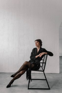 woman in a chic black outfit sits elegantly on a contemporary chair in a simple indoor space. environment features textured white walls that enhance her stylish look.