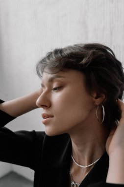 woman with short hair sits indoors, her head tilted slightly while she holds her hair back. She gazes thoughtfully to the side, surrounded by soft natural light and minimal decor.