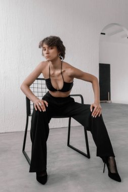 fashion model demonstrates confidence while seated on a minimalist black chair in a contemporary studio. She wears a black crop top and high-waisted pants, showcasing her bold style.