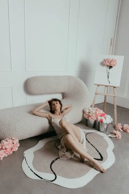 woman rests casually on a plush couch surrounded by beautiful flowers. scene has a modern aesthetic with art supplies nearby, capturing a peaceful moment.