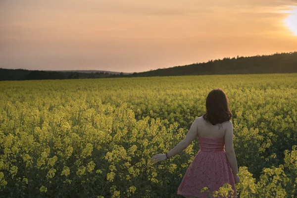 Attractive brunette girl in pink dress, walking through a yellow rapeseed culture enjoying the sunset light, in South Moravia region, Czech Republic.