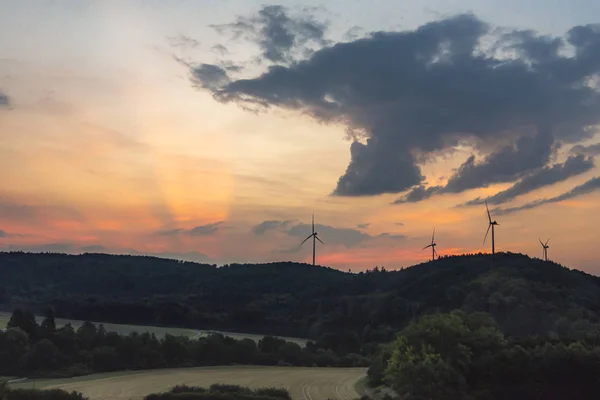 Beautiful sunrise landscape with wooded hills, meadows and modern wind turbines at the horizon, under a colorful sky, near Gaildorf town, Germany.