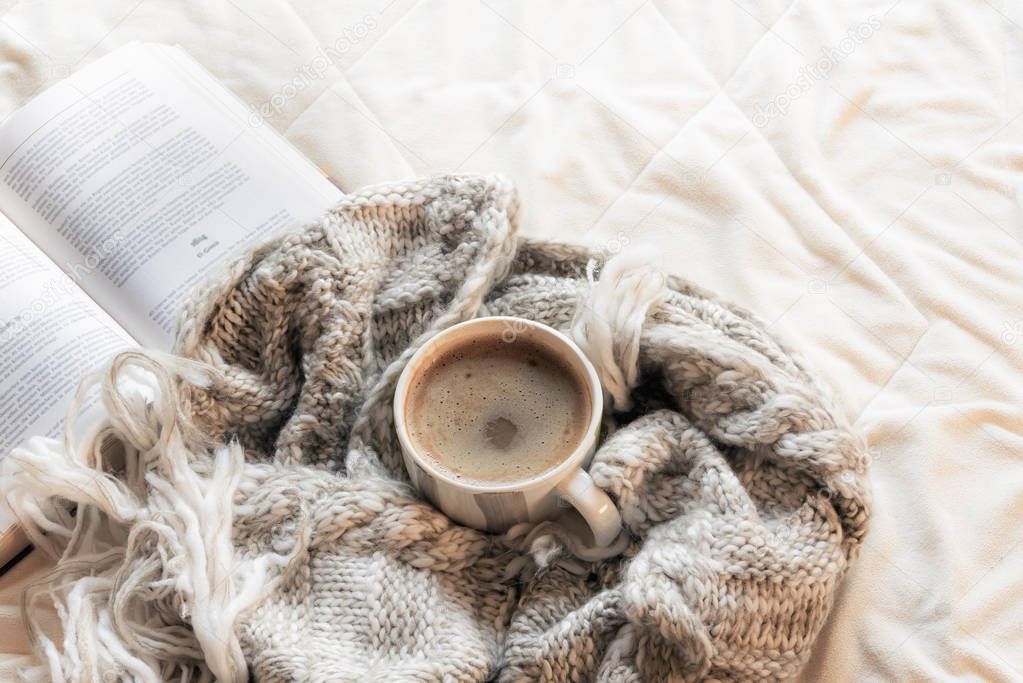 Fluffy shawl wrapped around a cup of hot chocolate, in front of an open book, on white sheets in a bright light, indoors.