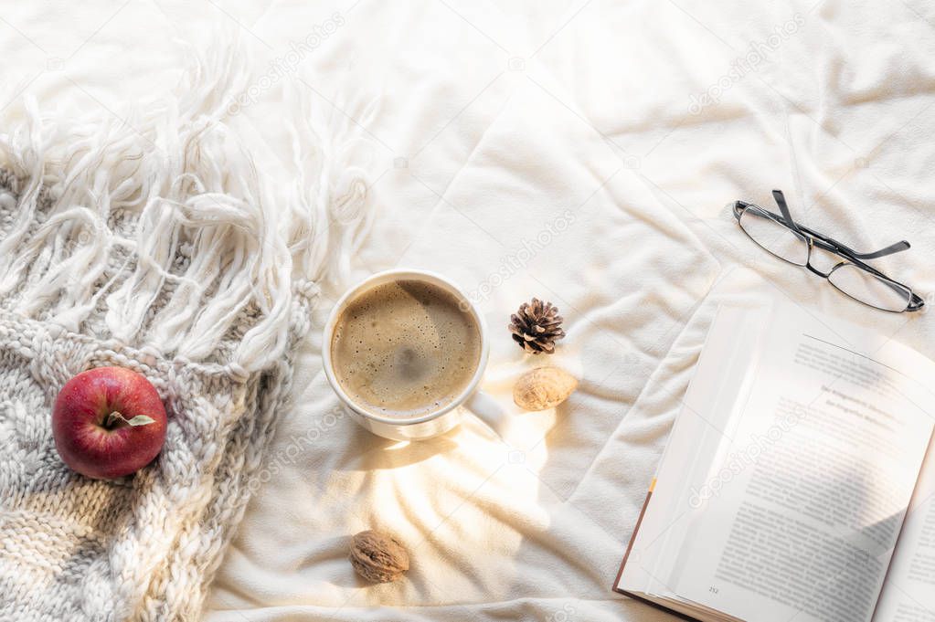 Cozy leisure concept with an open book, glasses, an apple on a fluffy shawl and a hot cup of coffee, on a white blanket, in the morning sun.