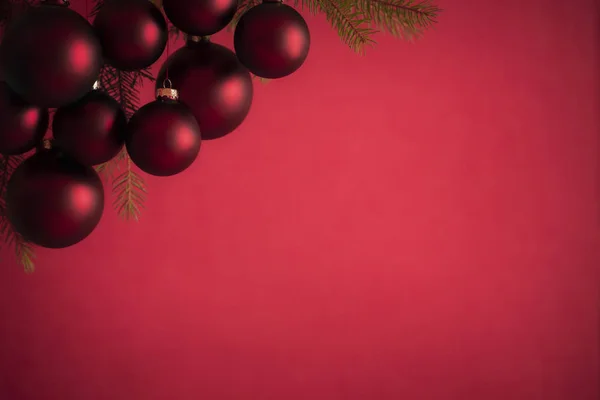 Dark red matte Christmas balls and fir twigs in the left corner on a bright red background. Perfect for a Christmas banner or greeting card.