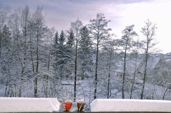 Two cups of hot coffee on a snow-covered railing from a balcony with the snowy trees in the background, on a sunny day of December