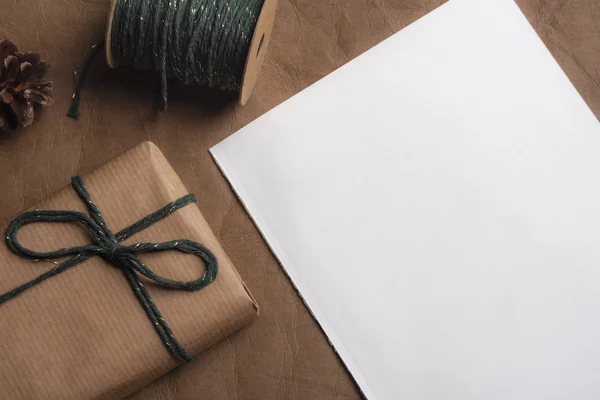Gift box wrapped in classic brown paper, tied with green and gold string and an unwritten white paper on a leather background.