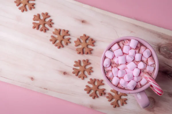 Winter sweets in pink shades, a cup of hot chocolate with mini marshmallows and a Christmas candy cane and snowflakes shaped gingerbread cookies.