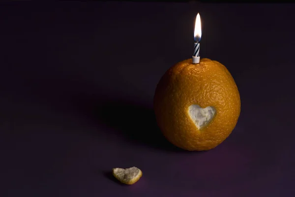 One orange with heart shaped peel cut of it and a lit birthday candle, on a purple background, in low light. Birthday greeting card.