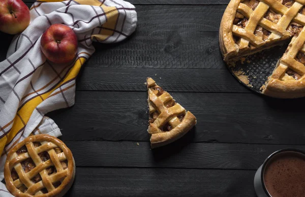 Rustic table with differently sized apple pies and one slice in the middle, a towel, apples, and hot chocolate. Above view. Traditional desserts.