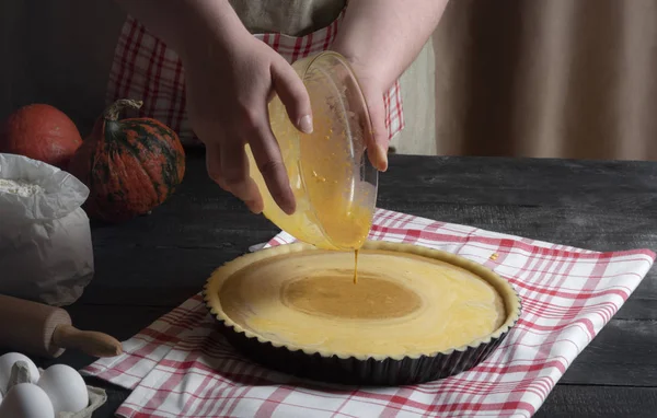 Woman hands pouring pie filling from a bowl into a tray with pastry dough, surrounded by ingredients for pumpkin pie. Classic sweet pie cooking.