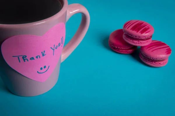 Pink coffee mug and macaroons on a blue background. Paper note with a thank you message fixed on the coffee cup