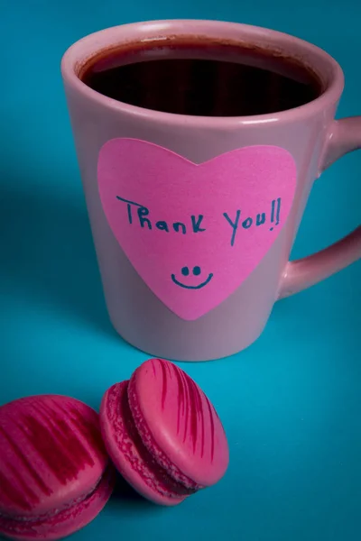 Thank you message handwritten on a heart-shaped sticky note. Pink mug of coffee and two pink macaroons on a blue table
