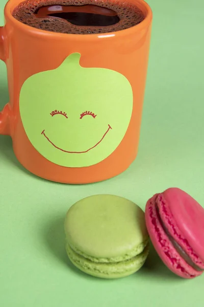 Cup of coffee with a sticky note on it and colorful macaroons on a green table. Smiley face drawn on the paper note