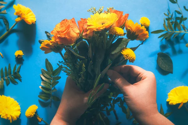 Woman hands making a flower bouquet from vibrant yellow and orange flowers over a blue paper background. Flat lay.