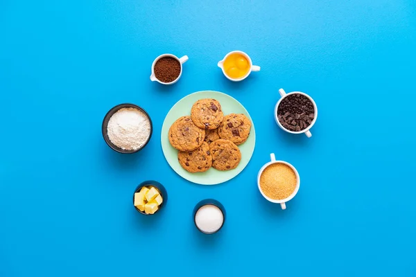 Chocolate chip cookies on a plate and the ingredients for making cookies, on a seamless blue background. Flat lay with recipe ingredients in bowls.