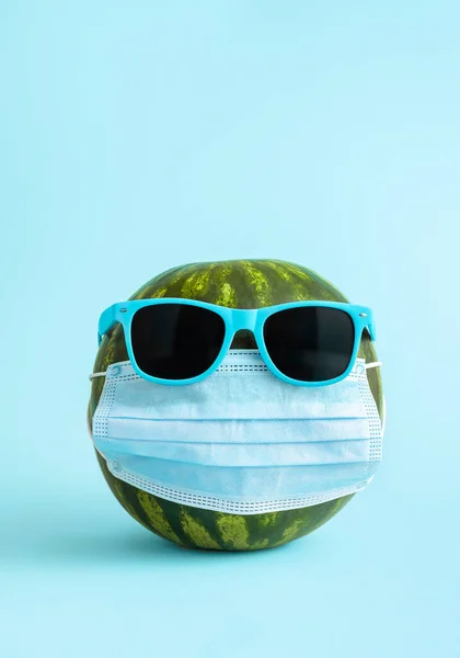 Watermelon with protective medical mask and sunglasses on blue background. Summer safety measures during the summer vacation concept. Coronavirus mask