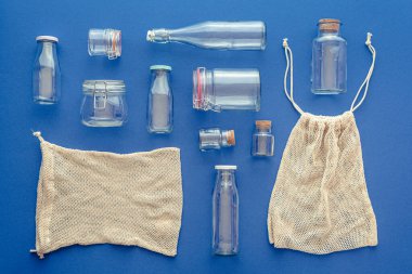 Yukarıda mavi arka planda cam kavanozlar ve şişelerle. Alışveriş torbaları için kullanılan eko poşetler. Ev mutfağı ve alışveriş için plastik olmayan konteynırlar.