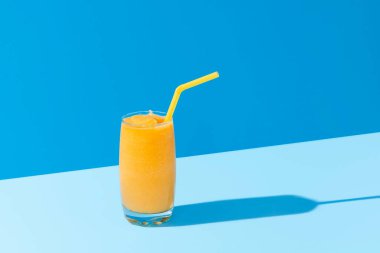 Sarı pipetli mango milkshake. Mavi arka planda, parlak ışıkta izole edilmiş. Bir bardak karışık mangolu smoothie. Yaz tropik kokteyli.