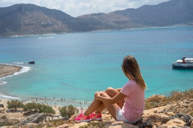 Büyük bir kayanın üzerinde oturan genç kadın ve mesafe içine deniz görünüyor. Seçici yumuşak odak.