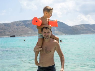 Mutlu baba veren omuz binmek plajda onun omuzlarında. Kameraya bakarak omuz baba üzerinde boy mutlu gülümseyerek. Baba ve oğul açık zevk. Mutlu yaz günlerinde. Balos lagün. Elafonisi Beach