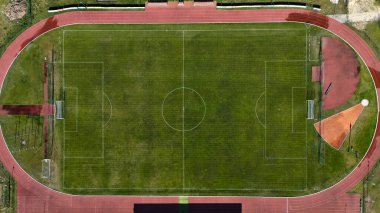 Kırmızı koşu pisti ve atletizm bölgeleriyle çevrili yeşil çim sahası olan bir futbol (futbol) stadyumunun yukarıdan aşağıya insansız hava aracı çekimi. İmaj, spor altyapısı, atletizm, eğlence ve kentsel planlama kavramlarını resmetmek için idealdir..