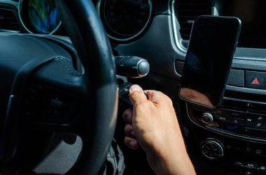 Bir erkeğin modern bir arabayı ateşleme anahtarı kullanarak çalıştırdığı yakın plan bir görüntü. Gösterge paneli, akıllı telefon montajı ve direksiyon görünür durumda. Araç işlemini gösteren yüksek çözünürlüklü fotoğraf