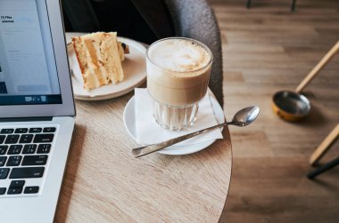 Laptop kek ve bir fincan kahveyle birlikte bir masanın üzerinde. Fincanın yanındaki masada bir kaşık var.
