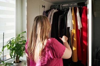 Kadın kıyafetlerle dolu dolabını karıştırıyor. Pembe bir gömlek giyiyor ve sarı saçları var.