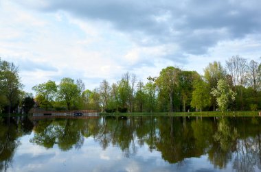 Arka planda ağaçlar ve bulutlu bir gökyüzü olan göl. Göl sakin ve huzurlu, ağaçlar suya yansıyor.