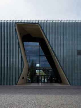 Binanın çok modern ve şık bir tasarımı var. Giriş, binaya açıklık ve şeffaflık hissi veren büyük cam bir pencereden oluşuyor. Bina bir şehirde yer almaktadır. Varşova 15.04.2025, Polonya