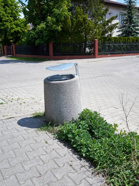 Çöp tenekesinin kapağı kaldırımda duruyor. Kapak açık ve çöp kutusu boş. Çöp tenekesi bir çitin ve ağacın yanında.