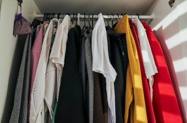 Dolapta kırmızı bir kazak ve beyaz bir gömlek var. Giysiler askıda asılı duruyor ve askıda da bir lavanta kesesi var. Sahne dağınık ve dağınık.