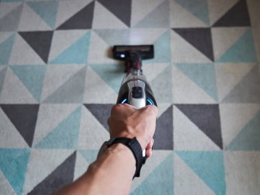 Üzerinde mavi düğme olan bir elektrikli süpürge tutuyor. Görüntünün rahat ve rahat bir havası var, çünkü kişi basit bir şekilde elektrikli süpürgeyi tutuyor ve acelesi yok.