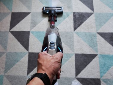 Üzerinde mavi düğme olan bir elektrikli süpürge tutuyor. Görüntünün rahat ve rahat bir havası var, çünkü kişi basit bir şekilde elektrikli süpürgeyi tutuyor ve acelesi yok.