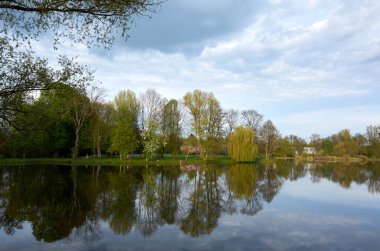 Arka planda ağaçlar ve bulutlu bir gökyüzü olan göl. Göl sakin ve huzurlu, ağaçlar suya yansıyor.