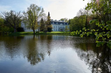 Büyük ev arka planında ağaçlar olan bir göl üzerinde yer alır. Su sakin ve gökyüzü berrak. Radziejowichi Sarayı 21.04.2025 Polonya