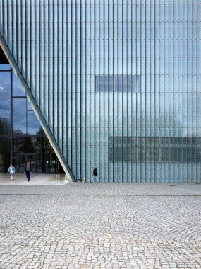 Adam köpeğiyle büyük bir cam binanın önünde yürüyor. Bina modern bir tasarıma sahiptir ve camdan yapılmıştır. Sahne sakin ve sakin, adamla birlikte. Varşova 15.04.2025, Polonya