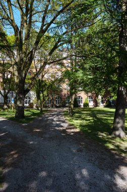 Ağaçlı bir parkta patika ve arka planda bir bina. Bina tuğladan yapılmış ve pencereleri var.