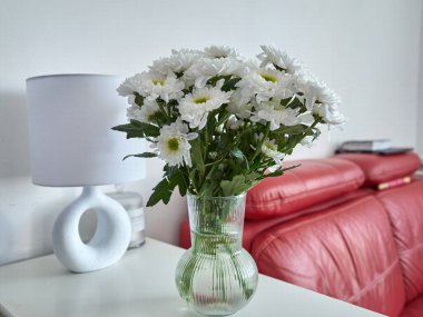 Vazo dolusu beyaz kasımpatı bir lambanın yanındaki masada duruyor. Çiçekler şeffaf bir vazoda ve onları bir buketin içindeymiş gibi gösterecek şekilde düzenlenmiş.