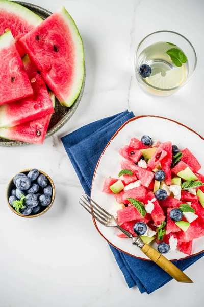 Summer food concept, fresh cold watermelon salad with feta cheese, blueberry, avocado and mint, white marble background copy space