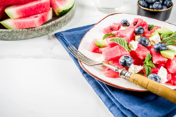 Summer food concept, fresh cold watermelon salad with feta cheese, blueberry, avocado and mint, white marble background copy space