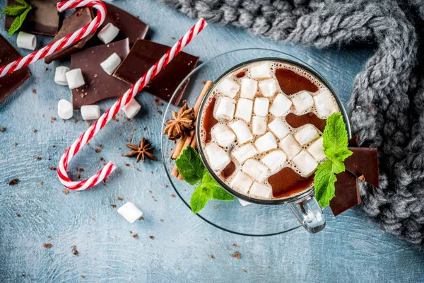 Homemade hot chocolate with mint, candy cane and marshmallow, light blue background with warm blanket, copy space