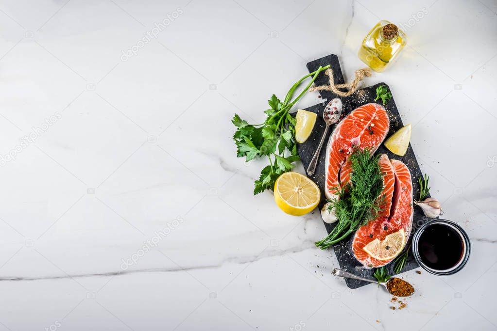 Raw salmon fish steaks with lemon, herbs, olive oil, ready for grill, slate cutting board, white marble background copy space above