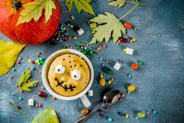 Kids Halloween food idea, funny mug cake decorate like monster face with marshmallow eyes and mouth painted with edible marker, blue background with sweets and autumn leaves, copy space