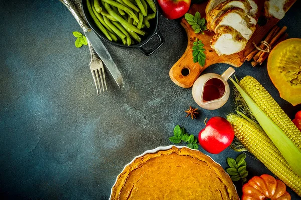 Thanksgiving holiday table with traditional festive food - turkey, pumpkin pie, pumpkins, green beans, cranberry sauce, corn, autumn apples, dark rusty table, top view space for text