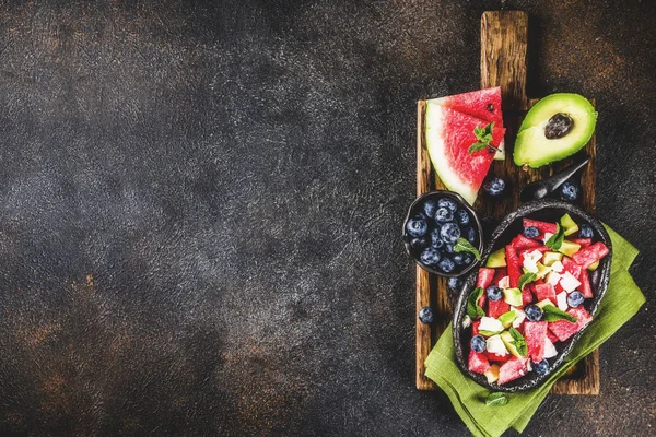 Summer food concept, fresh cold watermelon salad with feta cheese, blueberry, avocado and mint, dark rusty background copy space