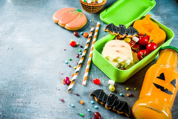 Halloween food, school lunch box with ghost sandwich, carved little pumpkins, cookies, candies and orange juice in funny monster bottle, blue concrete background top view copy space