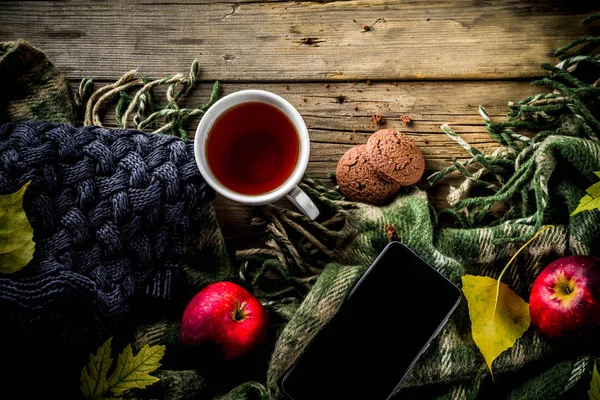 Autumn theme background, old wooden vintage planks boards, with plaid, sweater and autumn yellow leaves, tea cup, apples and chocolate chip cookies, with smartphone top view copy space